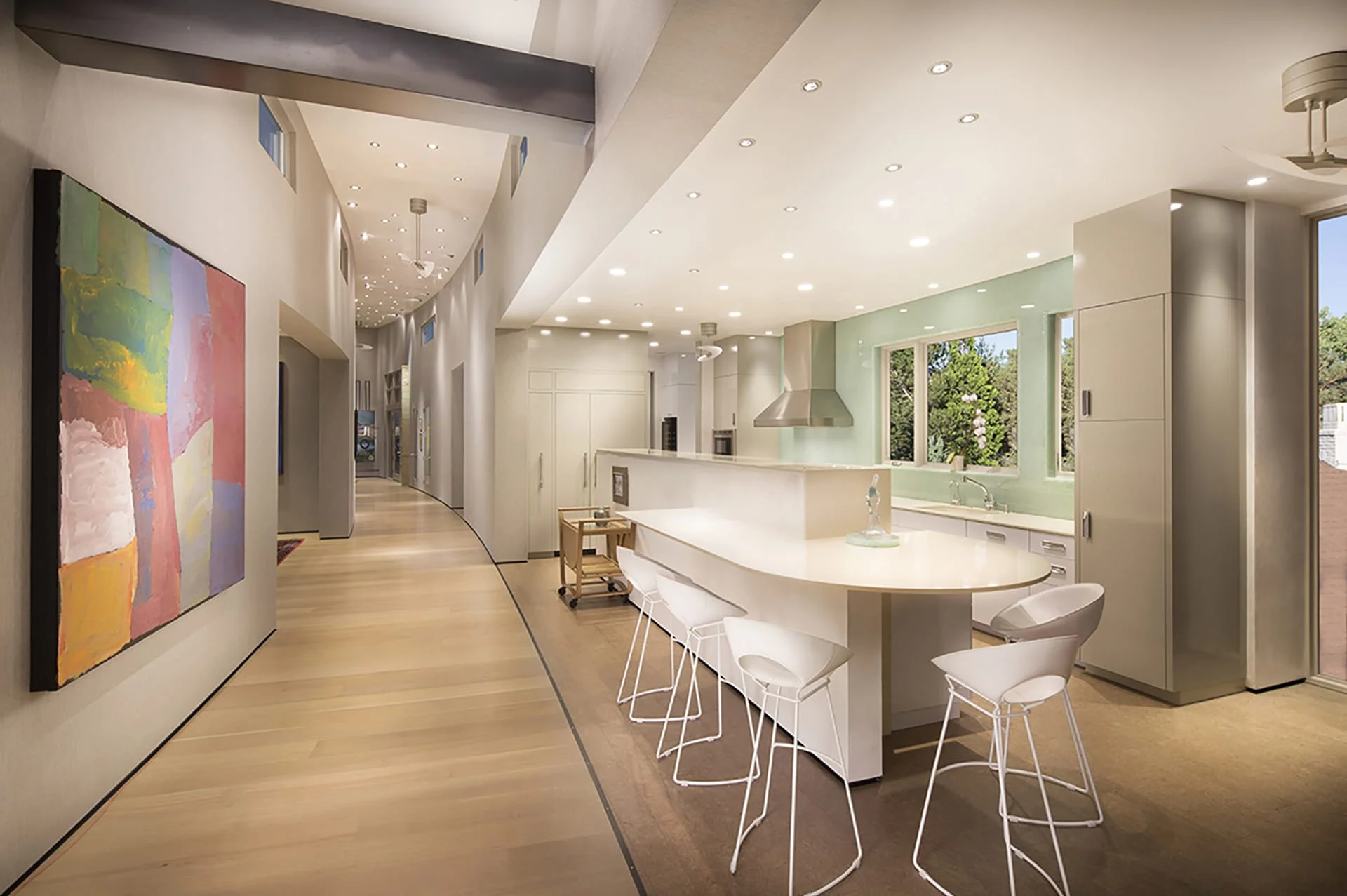 Curved hallway with recessed ceiling lights opening to a white kitchen and large painting