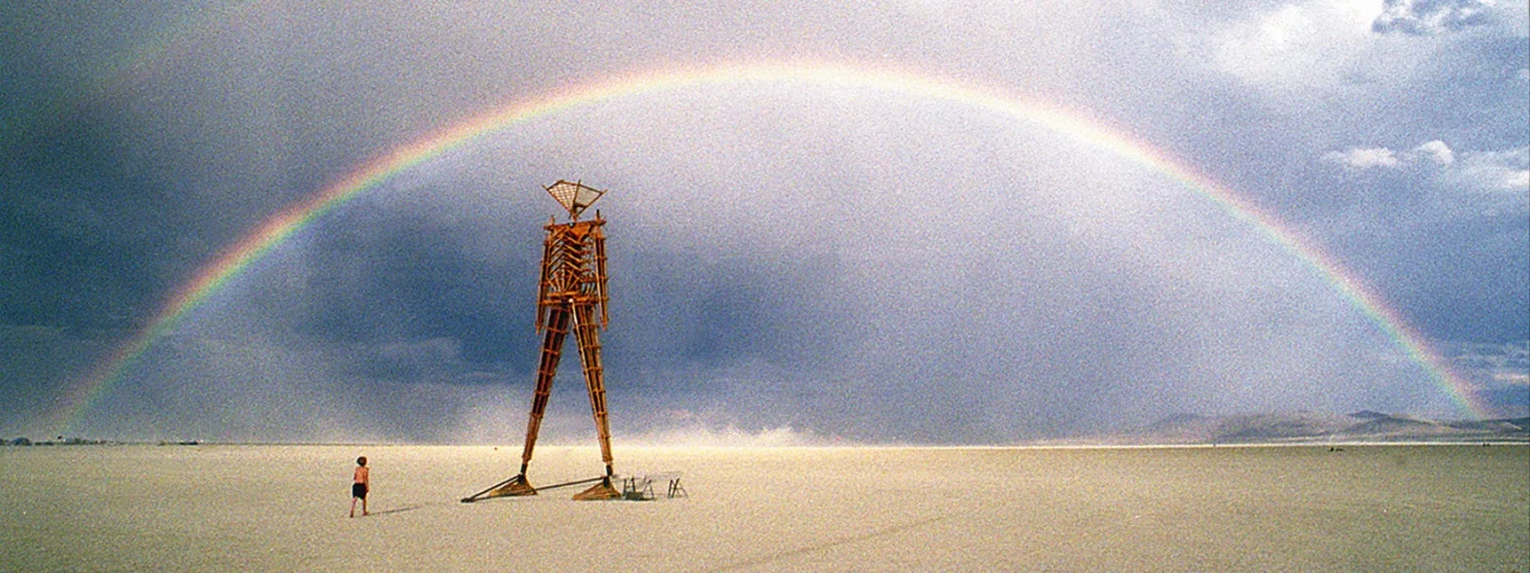 The Man standing on the open playa beneath a full rainbow