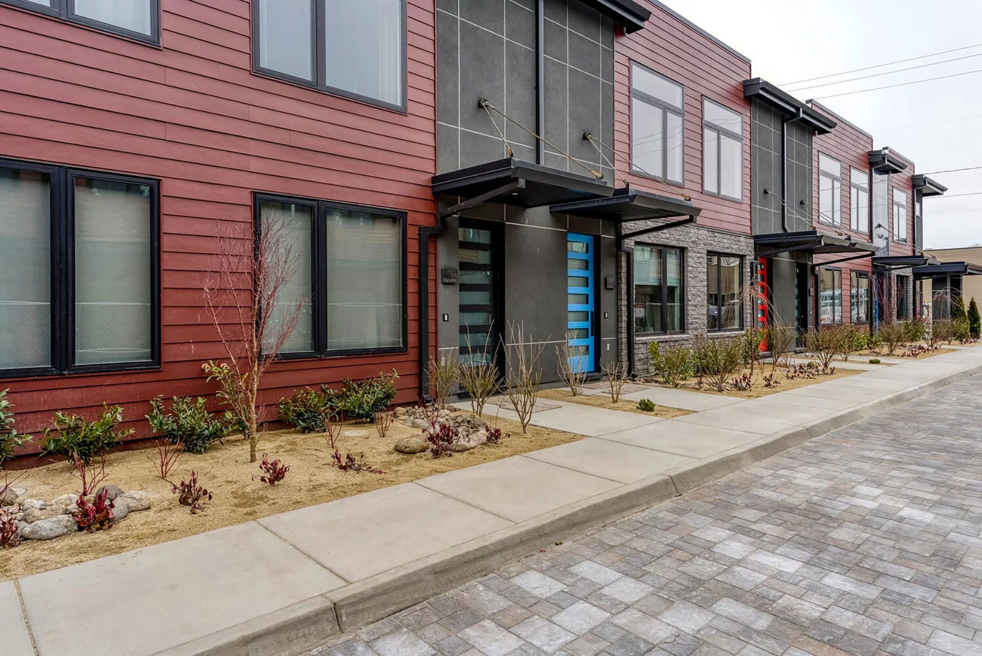 Row of townhomes with brick, horizontal siding, and steel panels along a paver street