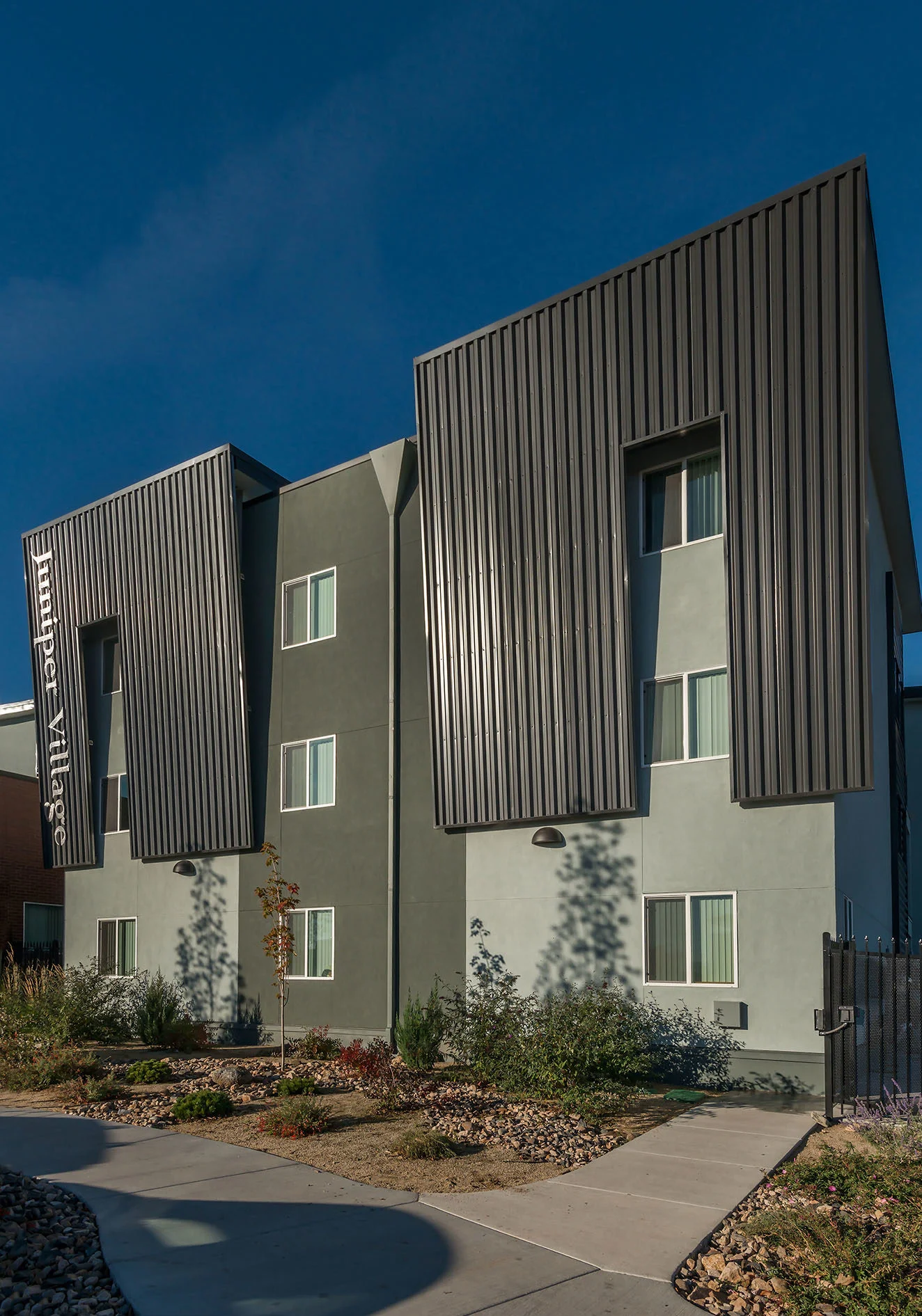 Dark corrugated metal panels folding off sage green walls along a landscaped walk