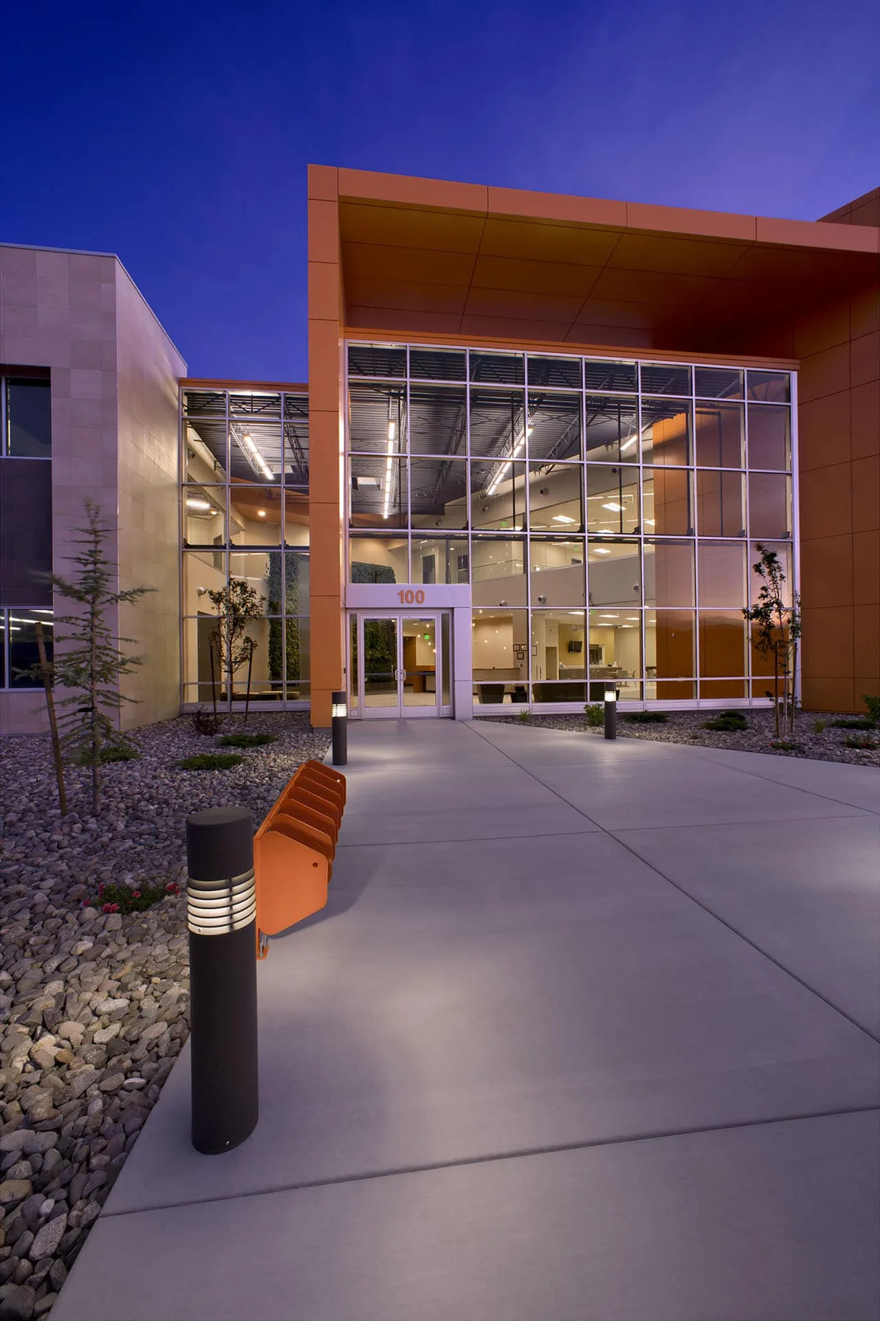 Glass entry framed by a burnt orange canopy wall, with lit bollards along the walk at dusk
