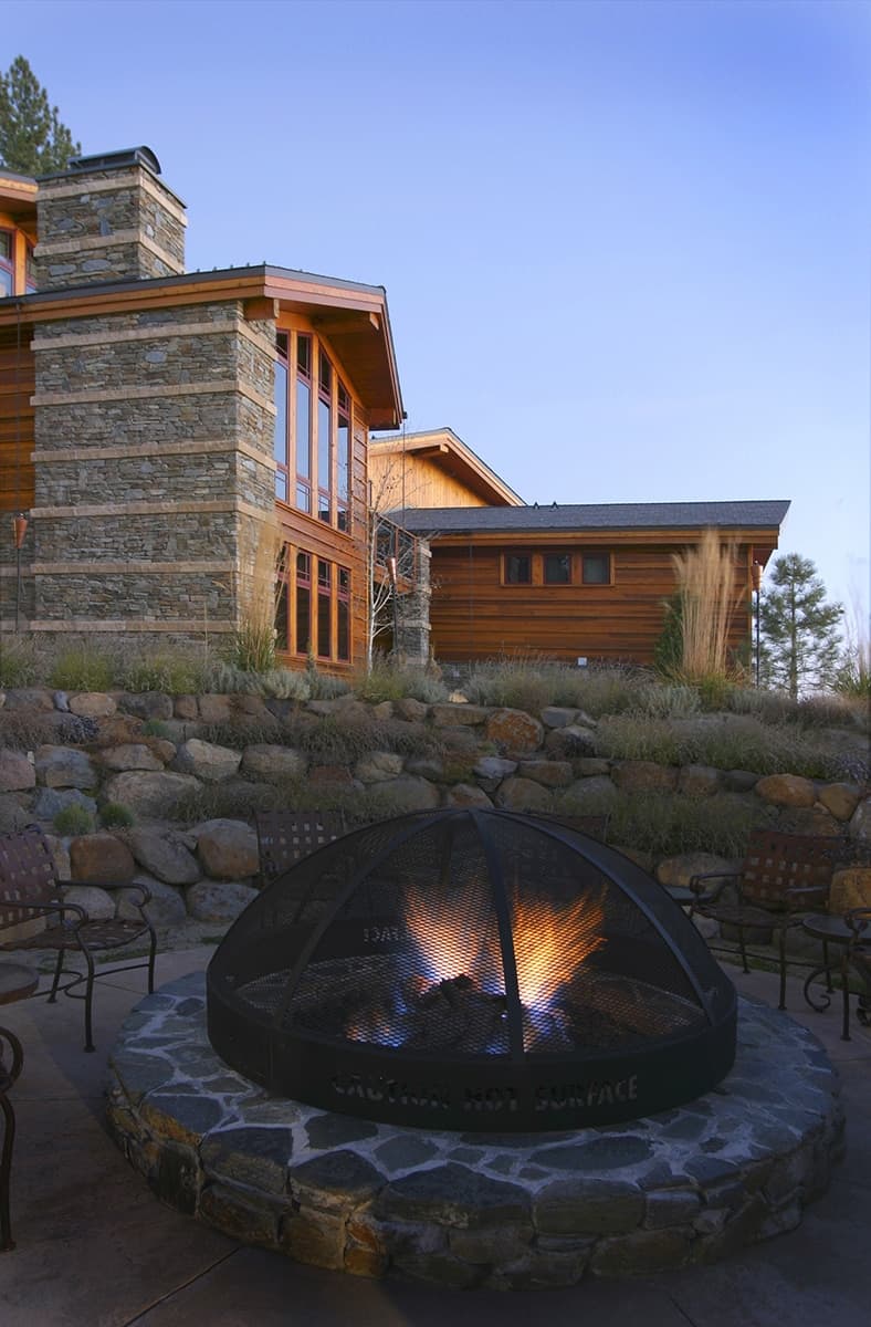 Stone-rimmed outdoor fire pit at dusk in front of a Tonopalo residence with stone chimney and cedar siding